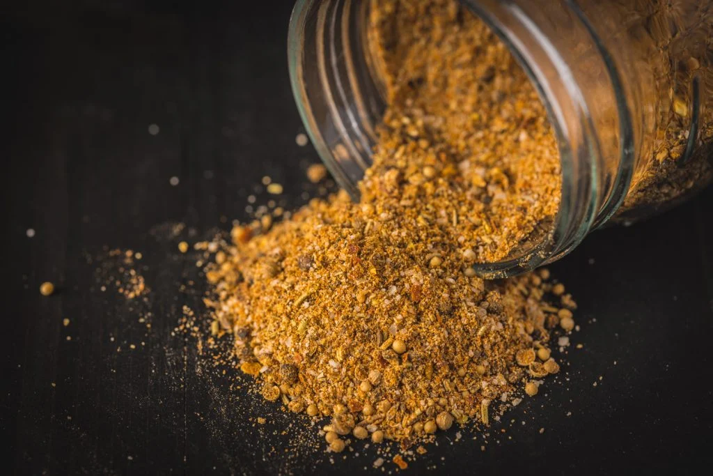 A small glass jar filled with homemade Montreal steak seasoning on a light kitchen counter, with scattered cracked pepper, garlic, and coriander seeds nearby