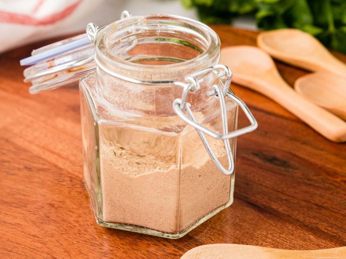 A small glass jar filled with homemade onion soup mix on a kitchen counter, with a spoon and small bowls of dried onion and spices nearby
