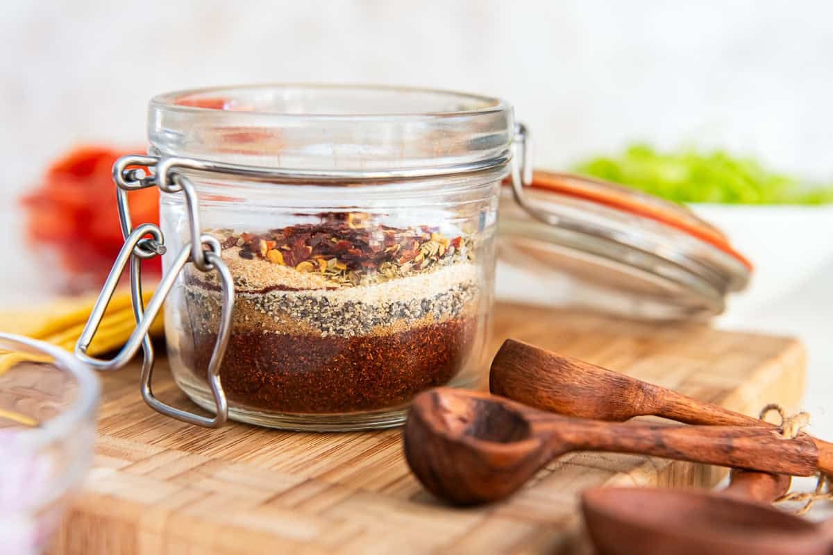 A small glass jar filled with homemade taco seasoning on a warm kitchen counter, with measuring spoons and bowls of spices nearby