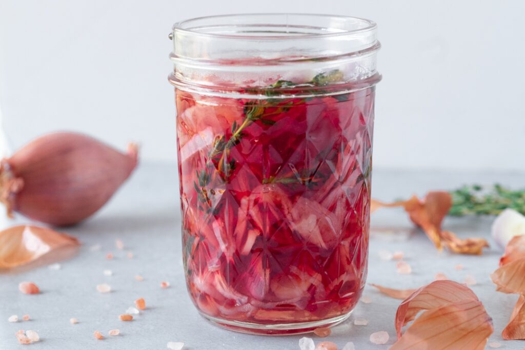 A small glass jar filled with pale pink quick pickled shallot rings in clear vinegar brine on a light kitchen counter, natural window light
