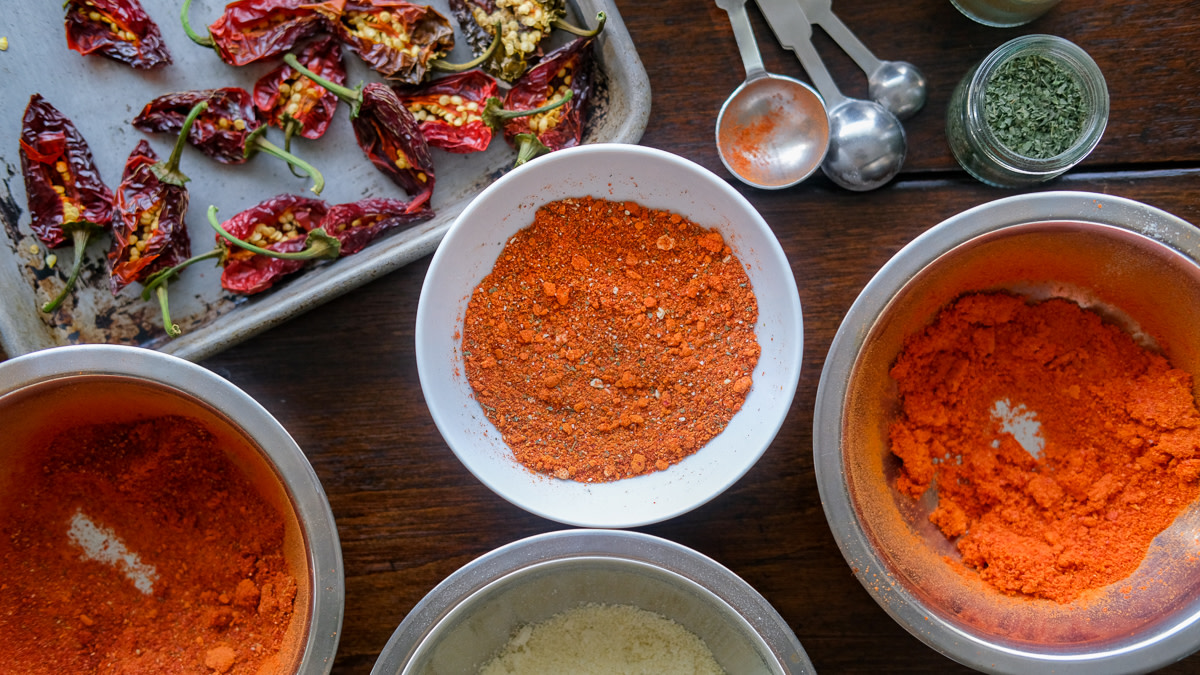 A small glass jar filled with reddish-brown homemade chili spice blend on a light kitchen counter, with measuring spoons and whole spices nearby