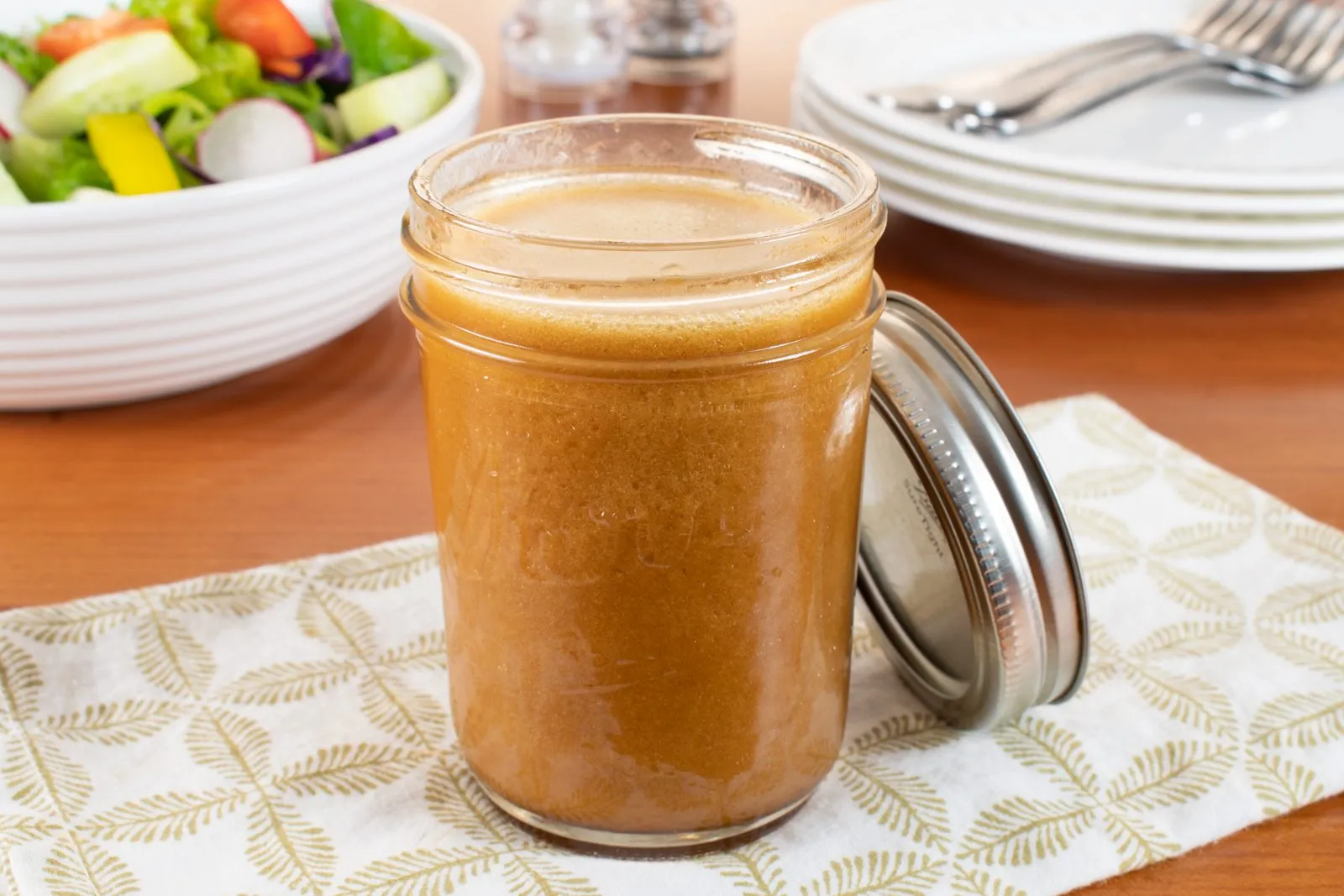 A small glass jar filled with sesame soy dressing with visible flecks of garlic and ginger, sitting on a kitchen counter next to a whisk