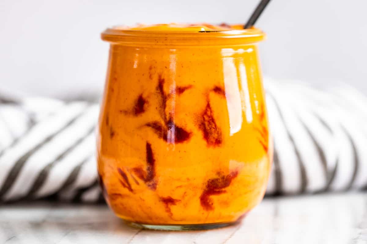 A small glass jar filled with sriracha mayo with the lid off on a kitchen counter next to a spoon, ready for storage in the refrigerator