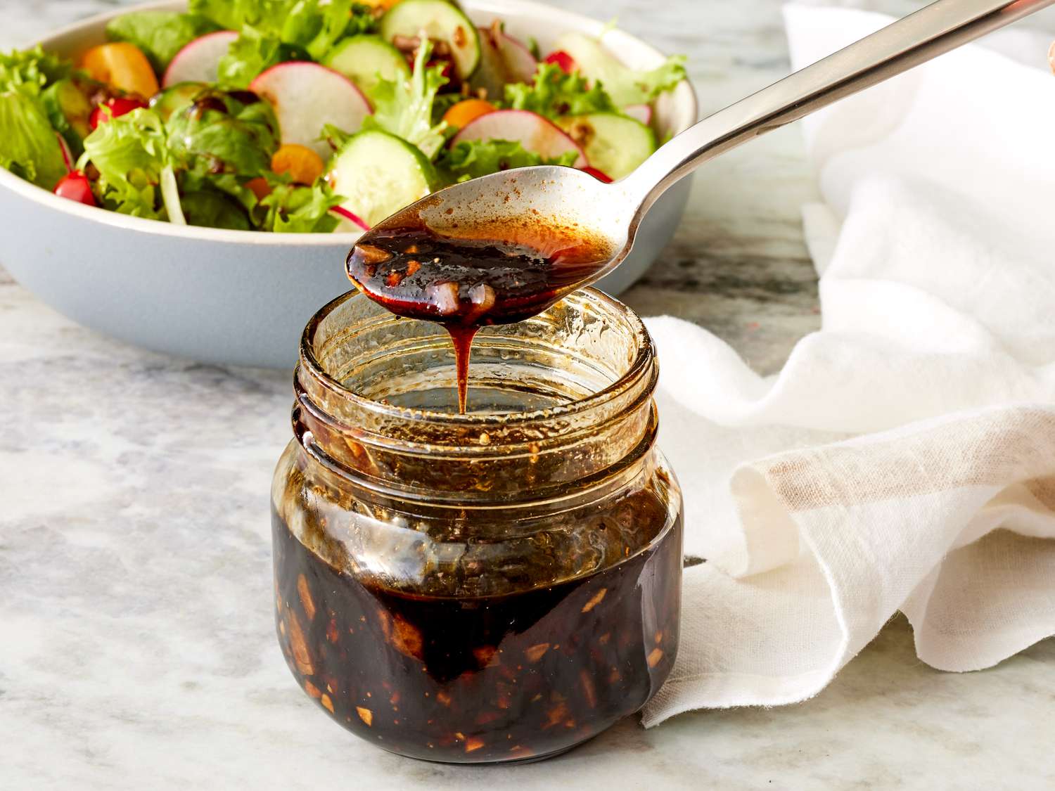 A small glass jar of balsamic vinaigrette with visible black pepper specks sitting on a bright kitchen counter beside a salad bowl and a whisk