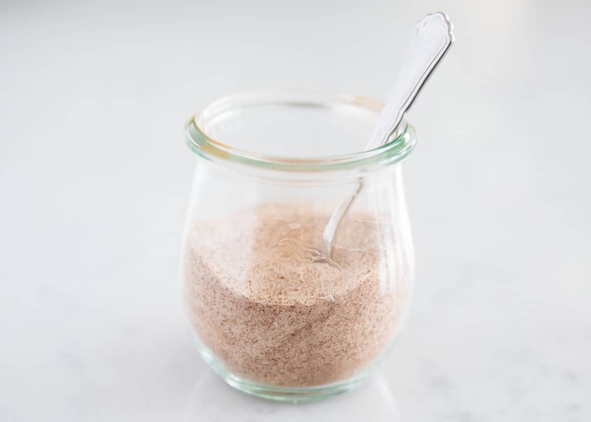 A small glass jar of cinnamon sugar on a warm-toned kitchen counter with a spoon and a few cinnamon sticks nearby