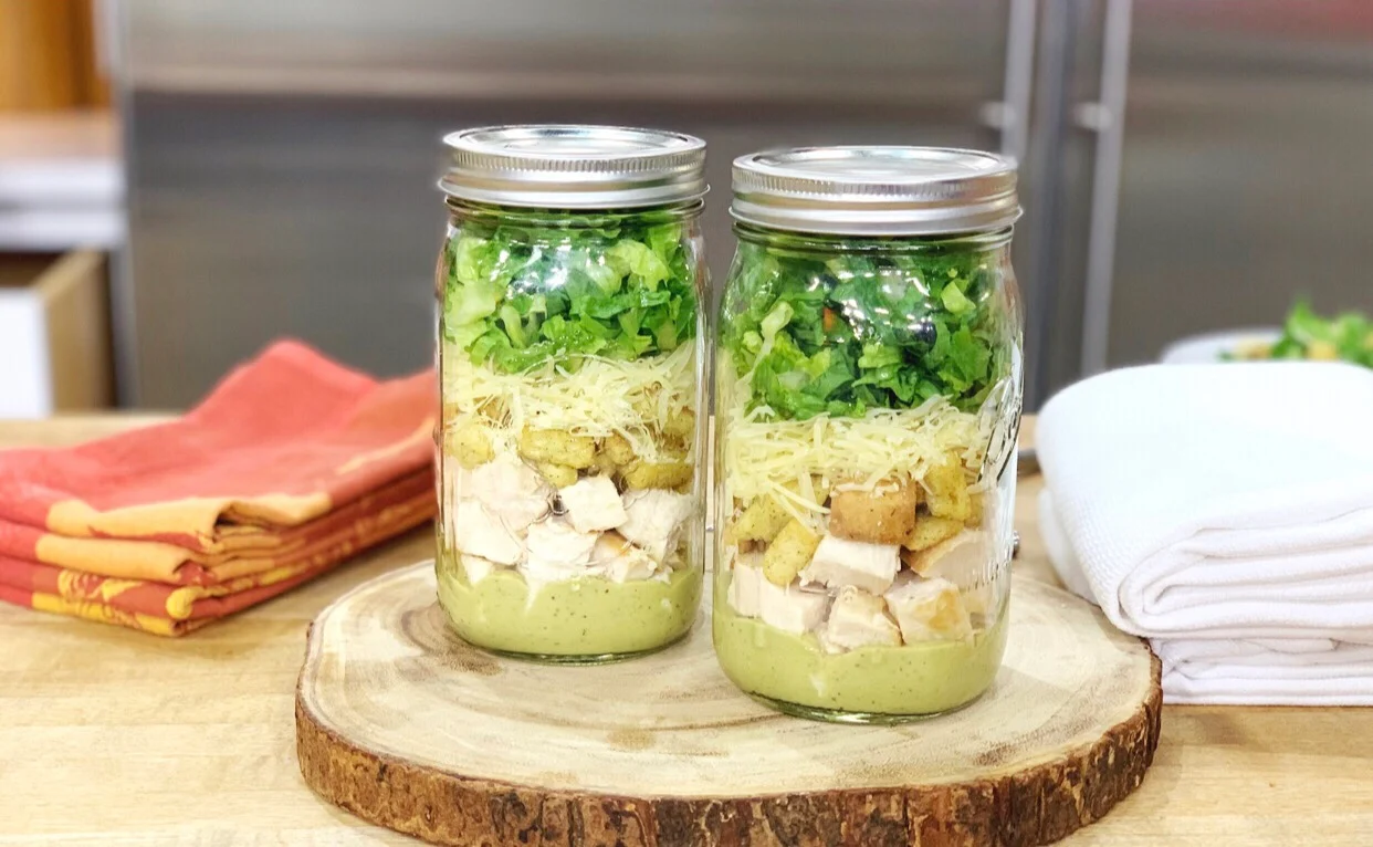 A small glass jar of creamy homemade Caesar dressing on a light kitchen counter with lemon wedges, a garlic clove, and shaved Parmesan nearby
