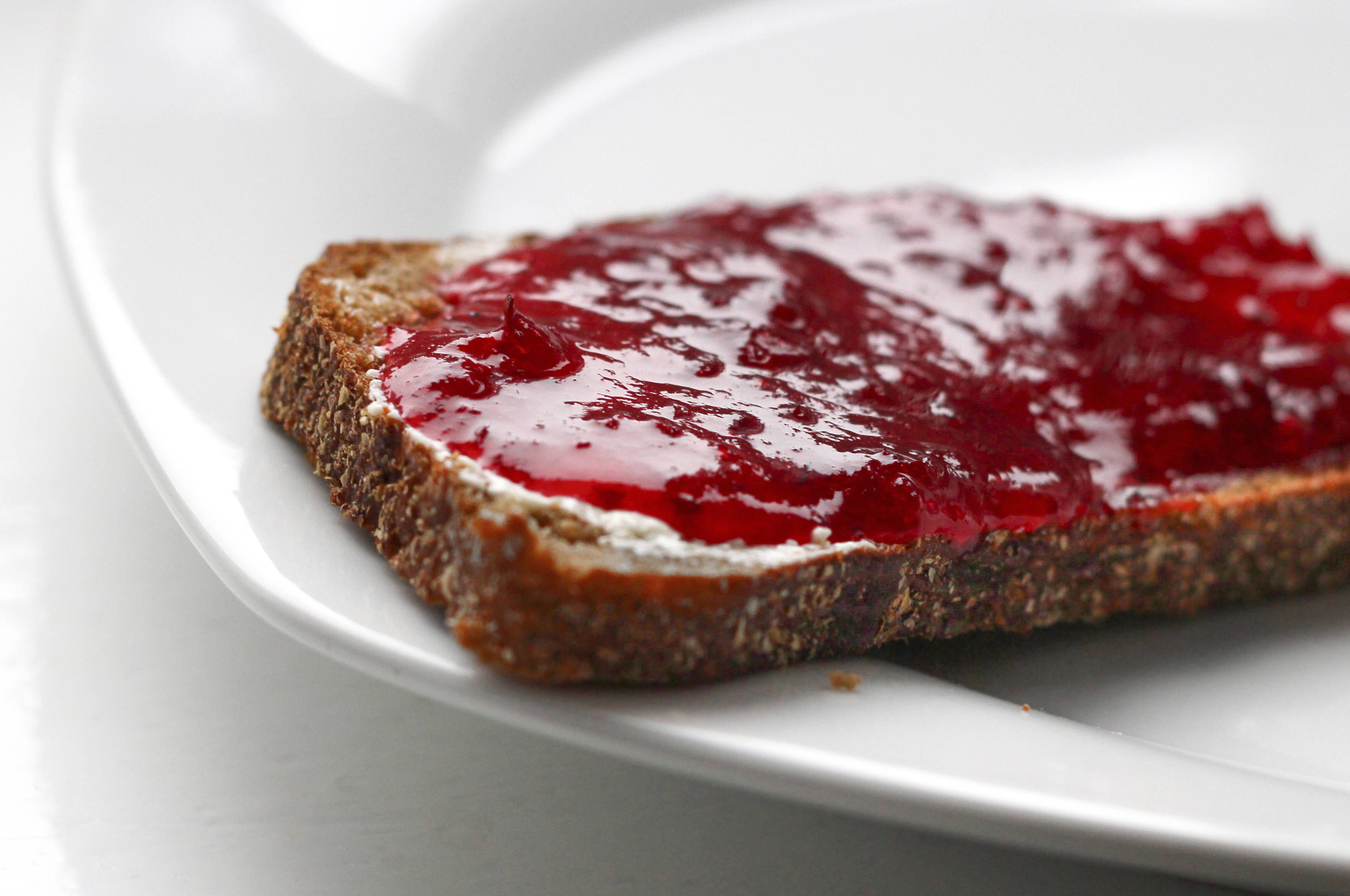 A small glass jar of deep purple muscadine jelly on a wooden table with a buttered slice of toast, a spoon, and a few whole muscadine grapes nearby in warm natural window light