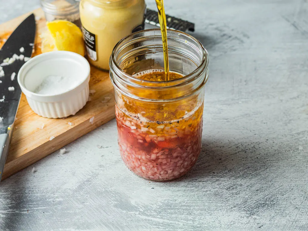 A small glass jar of deep ruby red wine vinaigrette on a light kitchen counter with a spoon nearby and a simple salad in the background, natural window light