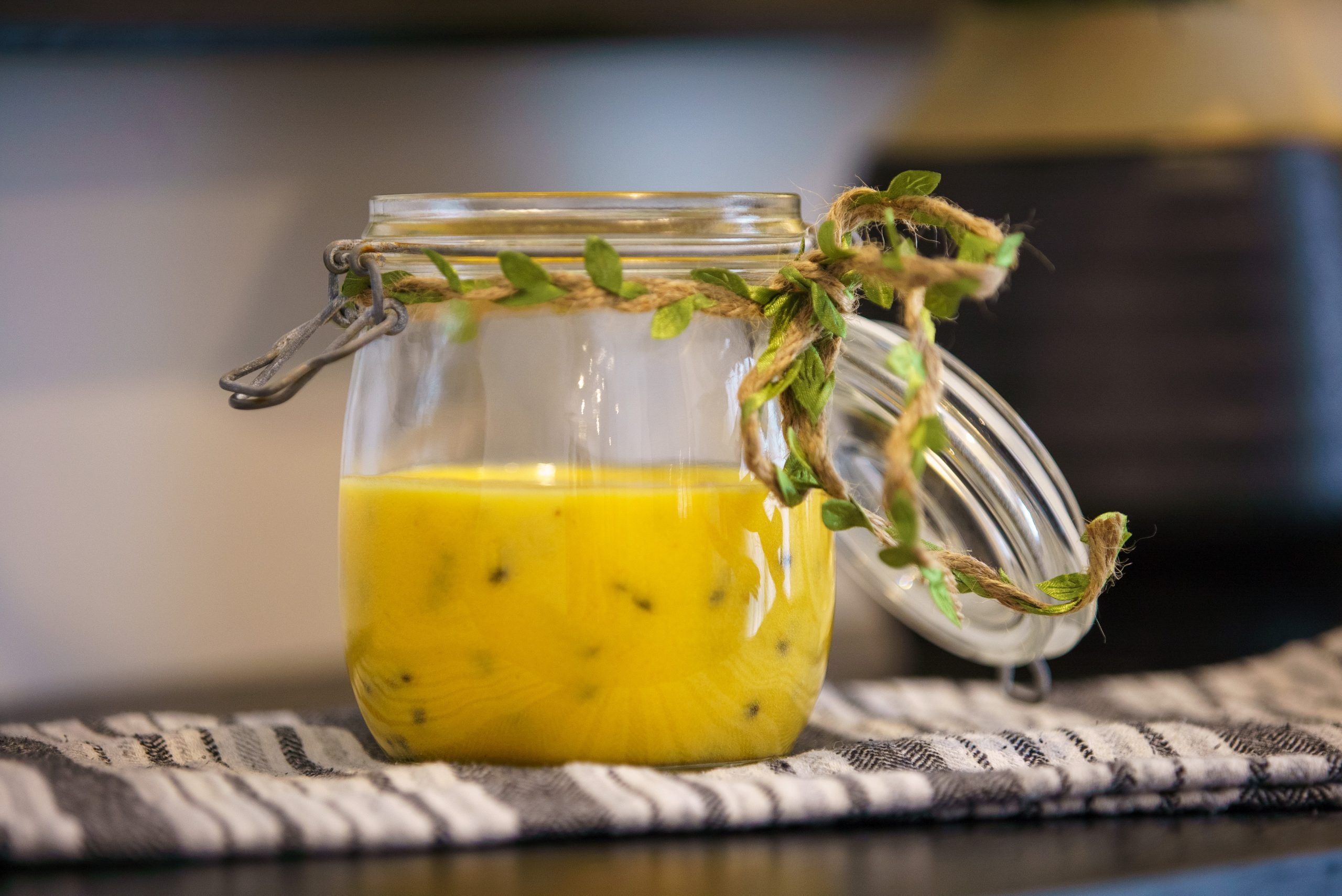 A small glass jar of glossy golden passionfruit curd on a light stone counter with a spoon resting beside it and fresh passionfruit halves nearby