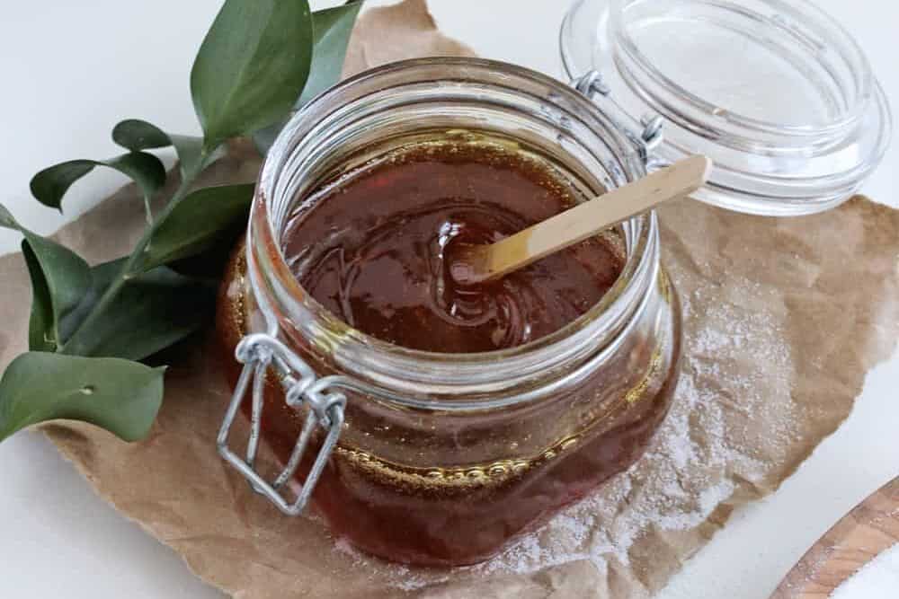 A small glass jar of golden homemade sugar wax on a warm toned kitchen counter with a wooden spatula beside it, natural window light