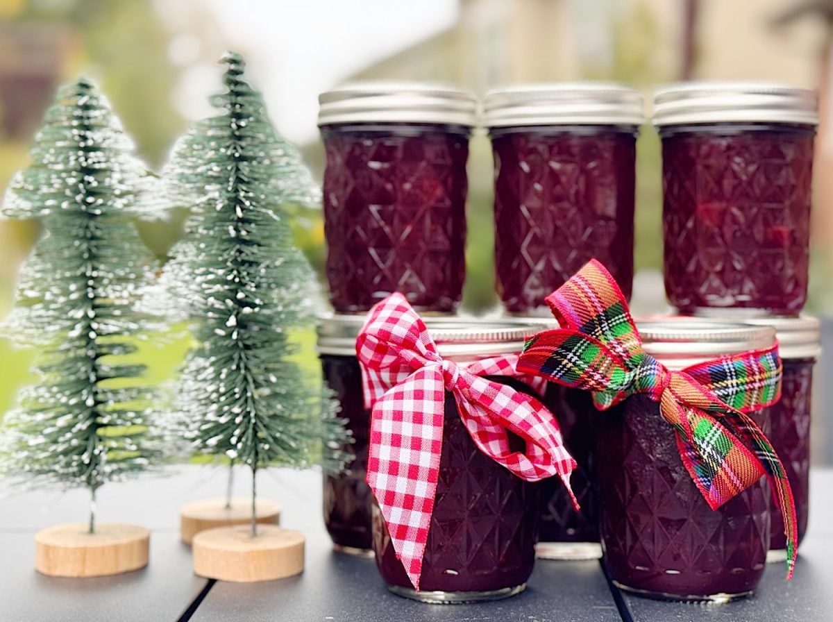 A small glass jar of homemade Christmas jam with cranberries and orange zest visible, sitting on a wooden kitchen counter with a spoon beside it and soft warm holiday lighting