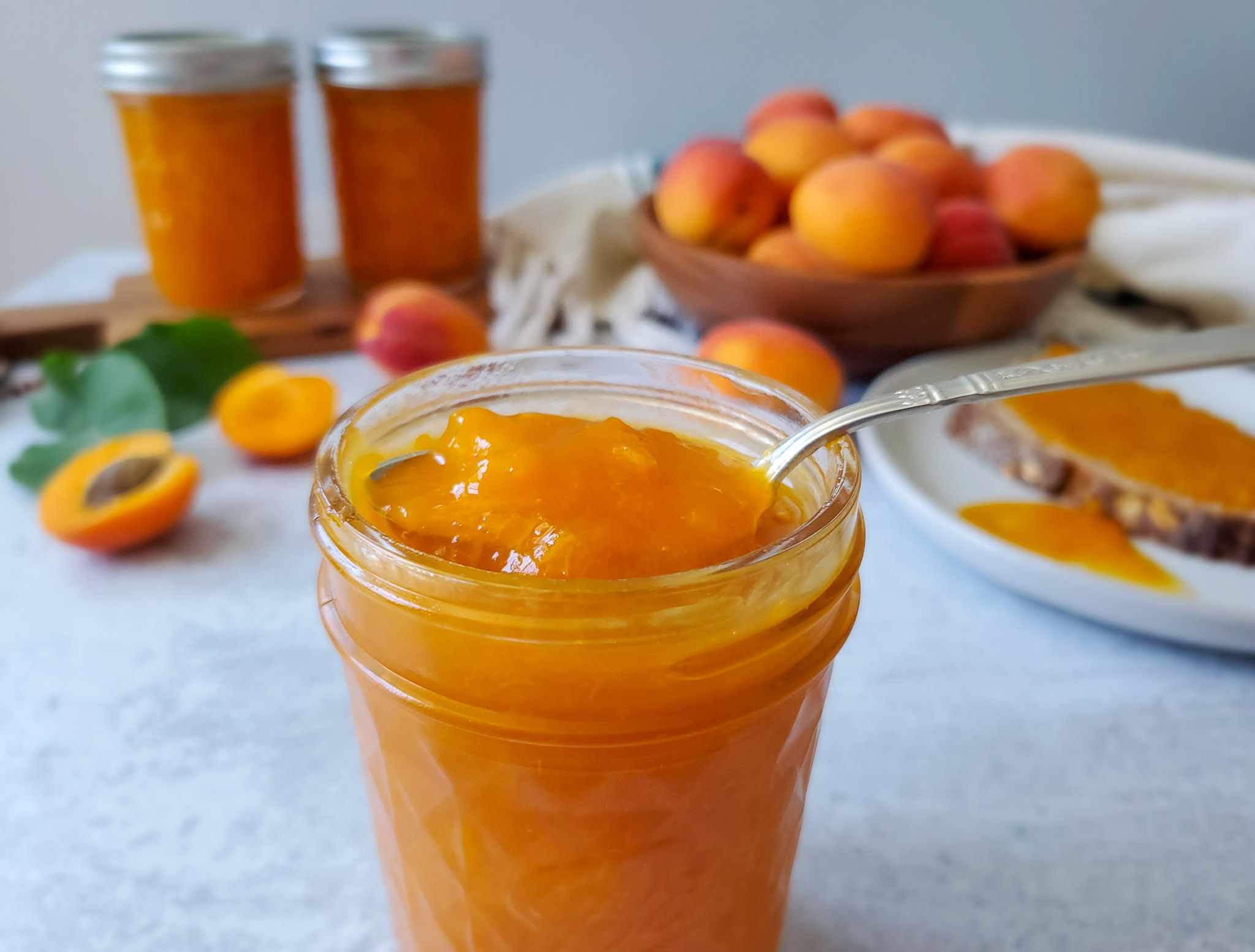 A small glass jar of homemade apricot jam with a spoon resting beside it on a warm-toned kitchen counter, natural window light