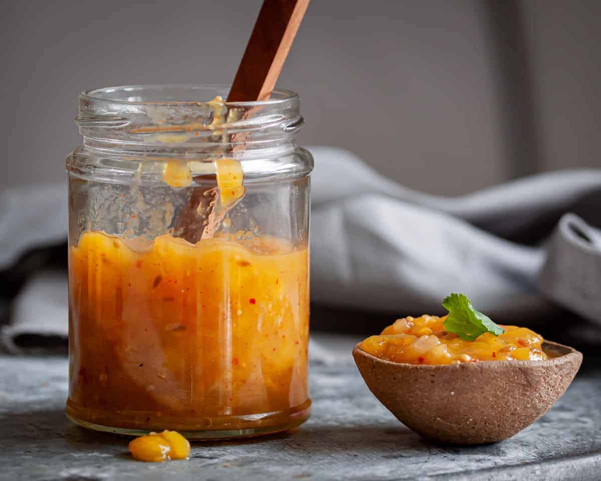 A small glass jar of homemade mango chutney with visible mango chunks and spices, sitting on a light stone kitchen counter beside a spoon
