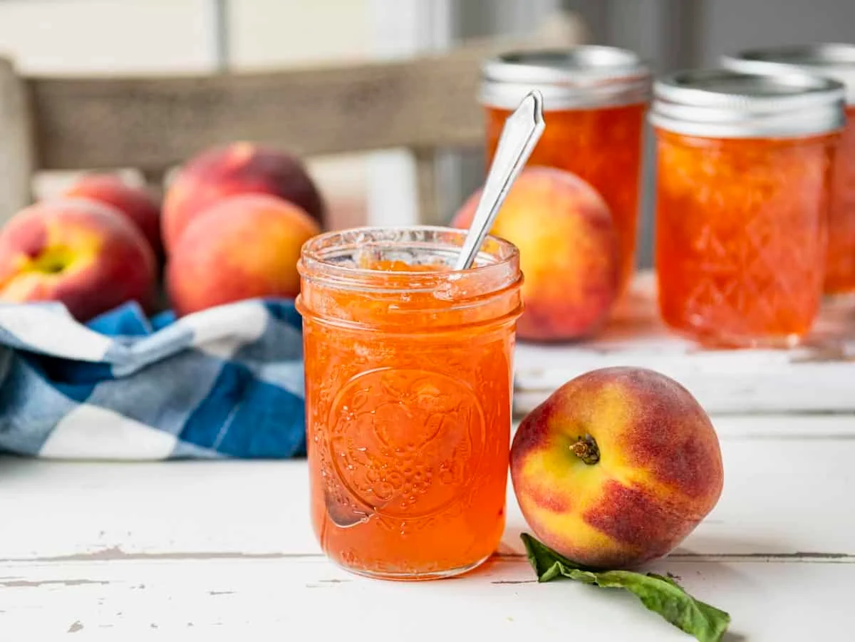 A small glass jar of homemade peach jelly with a glossy amber color on a wooden kitchen counter, peaches and a butter knife nearby in soft natural light