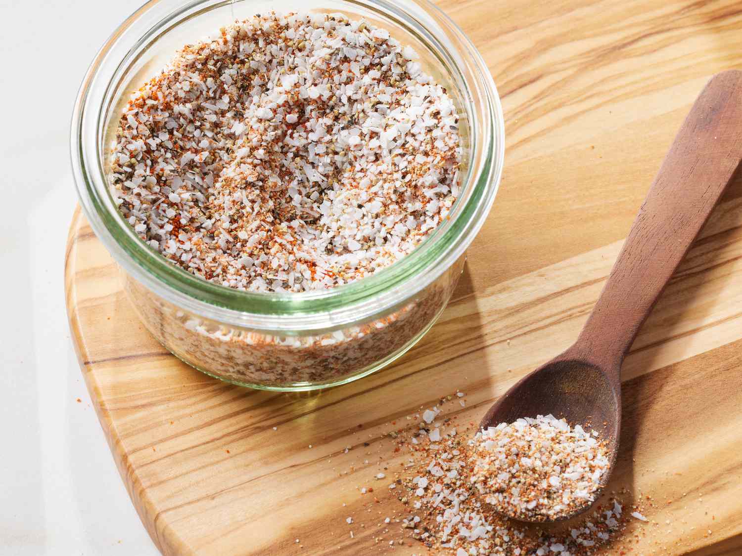 A small glass jar of homemade seasoned salt on a light kitchen counter with measuring spoons and a few spices nearby