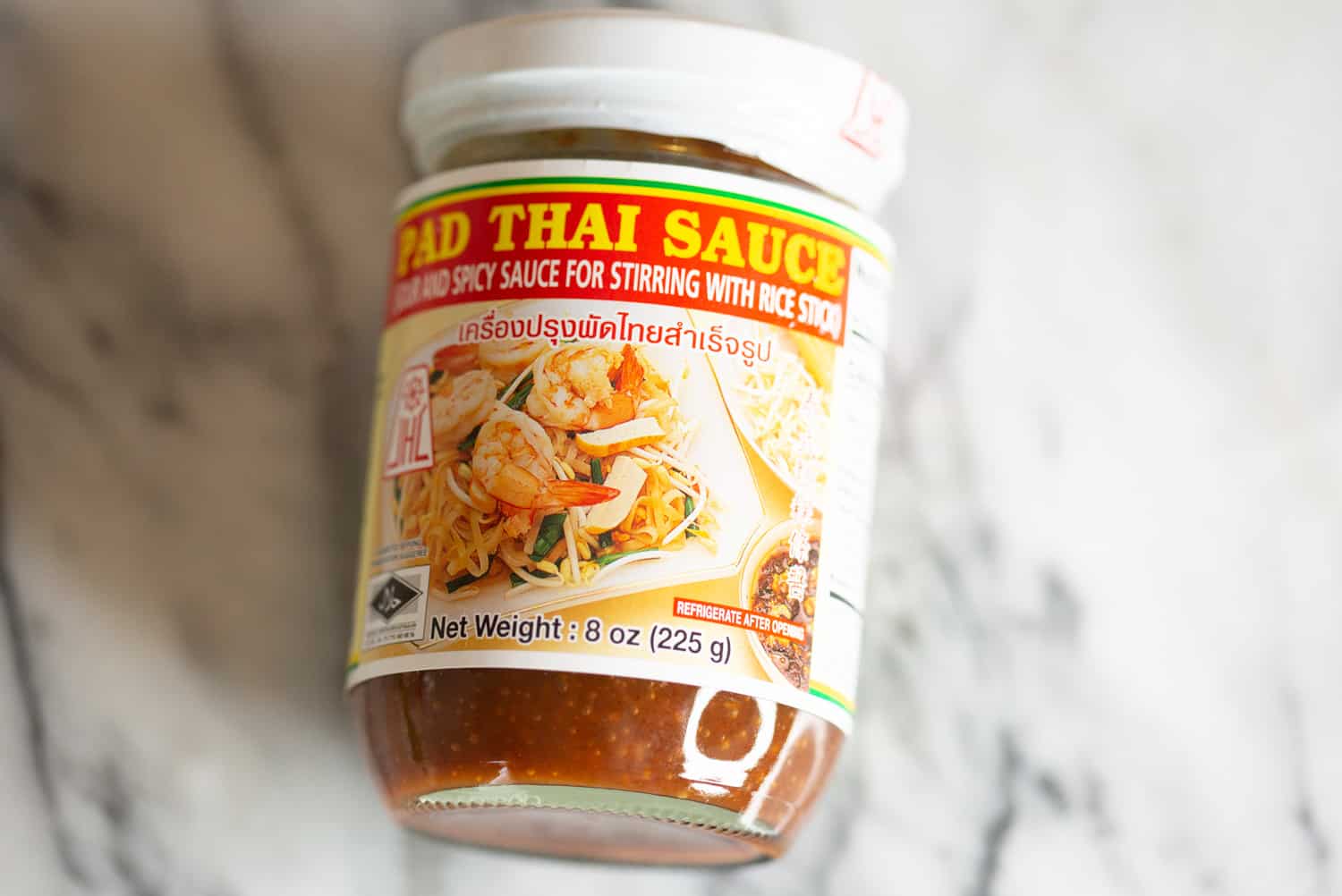 A small glass jar of pad thai sauce with a lid resting beside it on a refrigerator shelf next to fresh limes and herbs