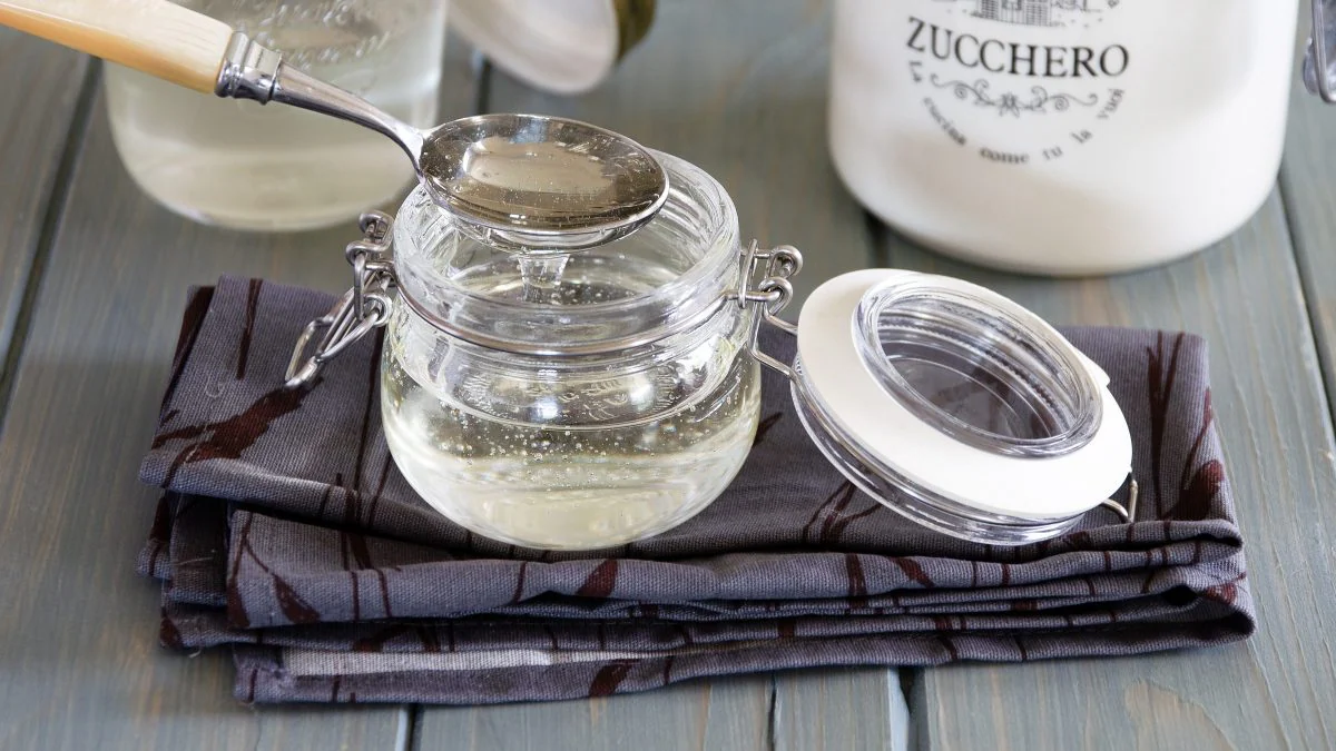 A small glass jar of simple syrup on a kitchen counter with a spoon nearby and a bowl of sugar behind it
