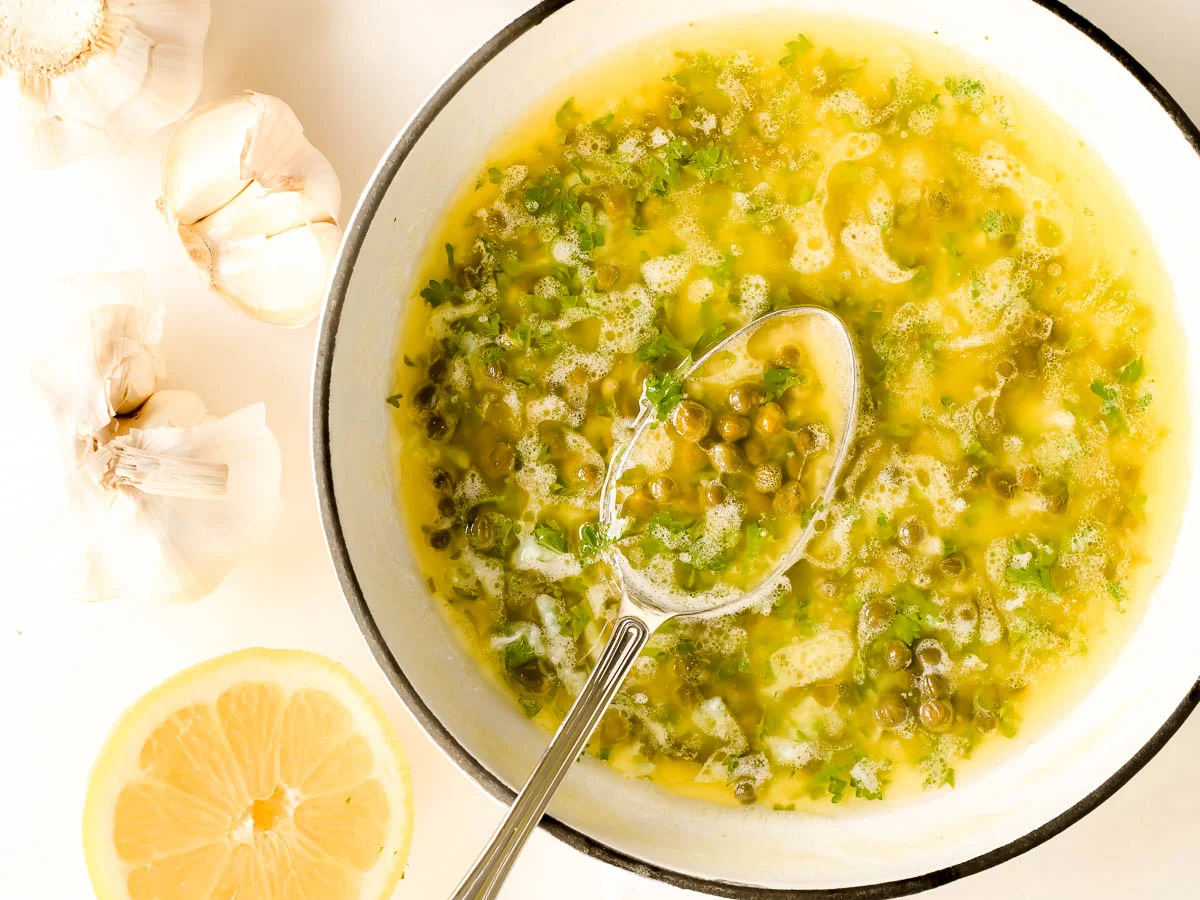 A small grouping of lemon, capers in a jar, garlic cloves, a stick of butter, and fresh parsley on a clean kitchen counter in natural window light