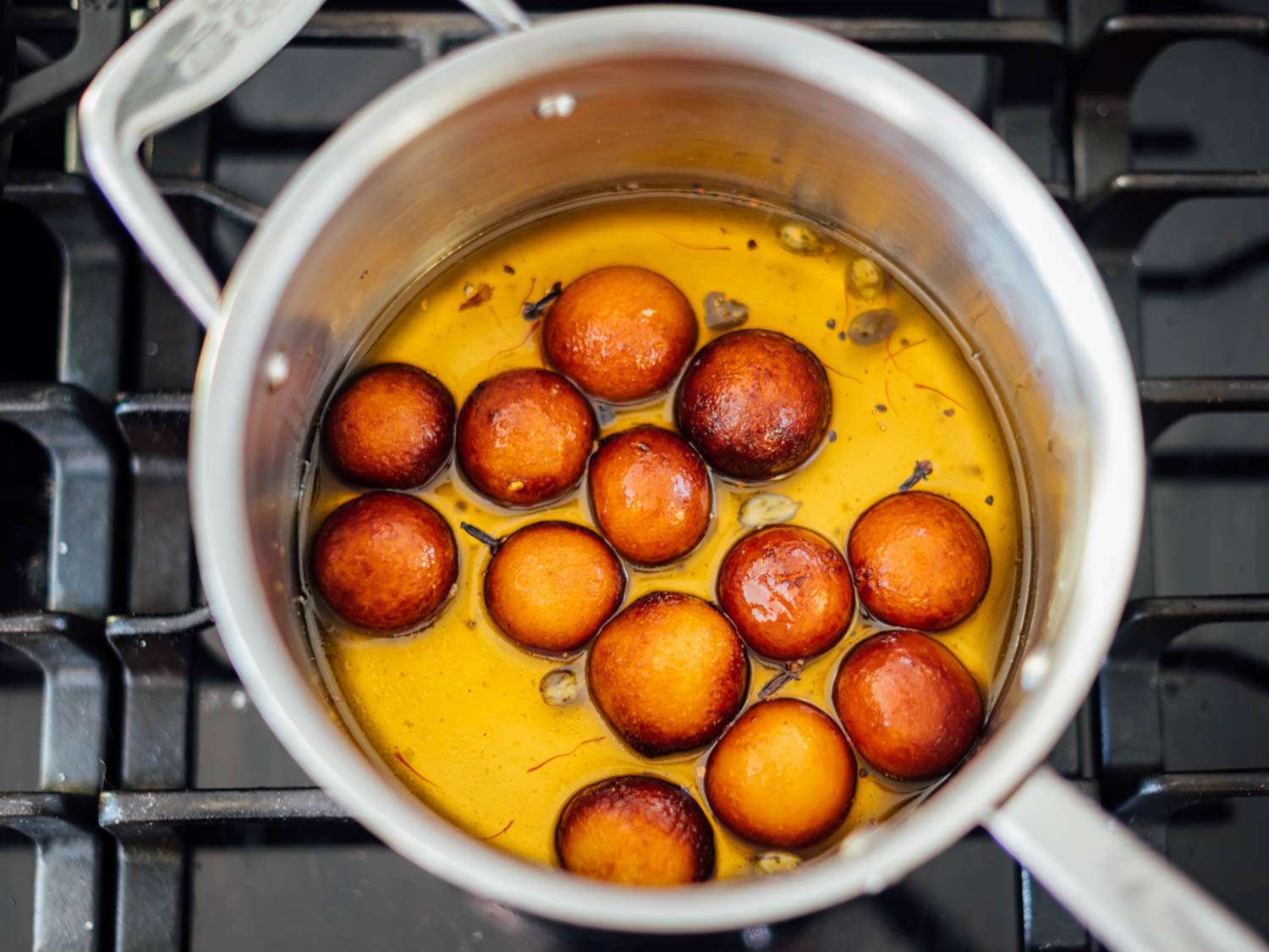A small saucepan filled with sugar syrup and a few gulab jamun floating on top, cardamom pods visible