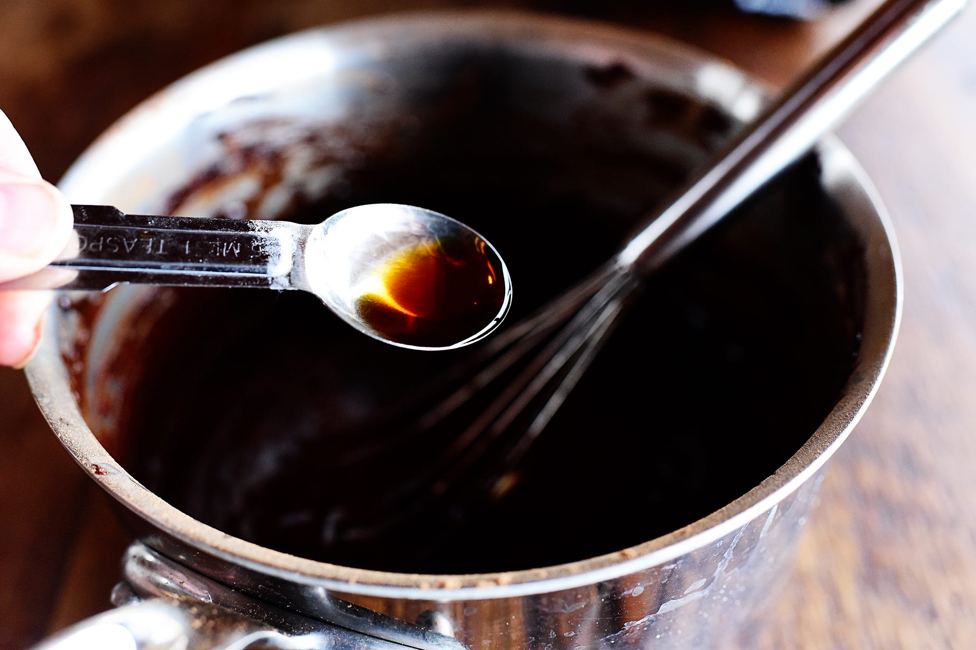 A small saucepan of glossy hot fudge sauce being stirred with a spoon on a stovetop, warm kitchen lighting