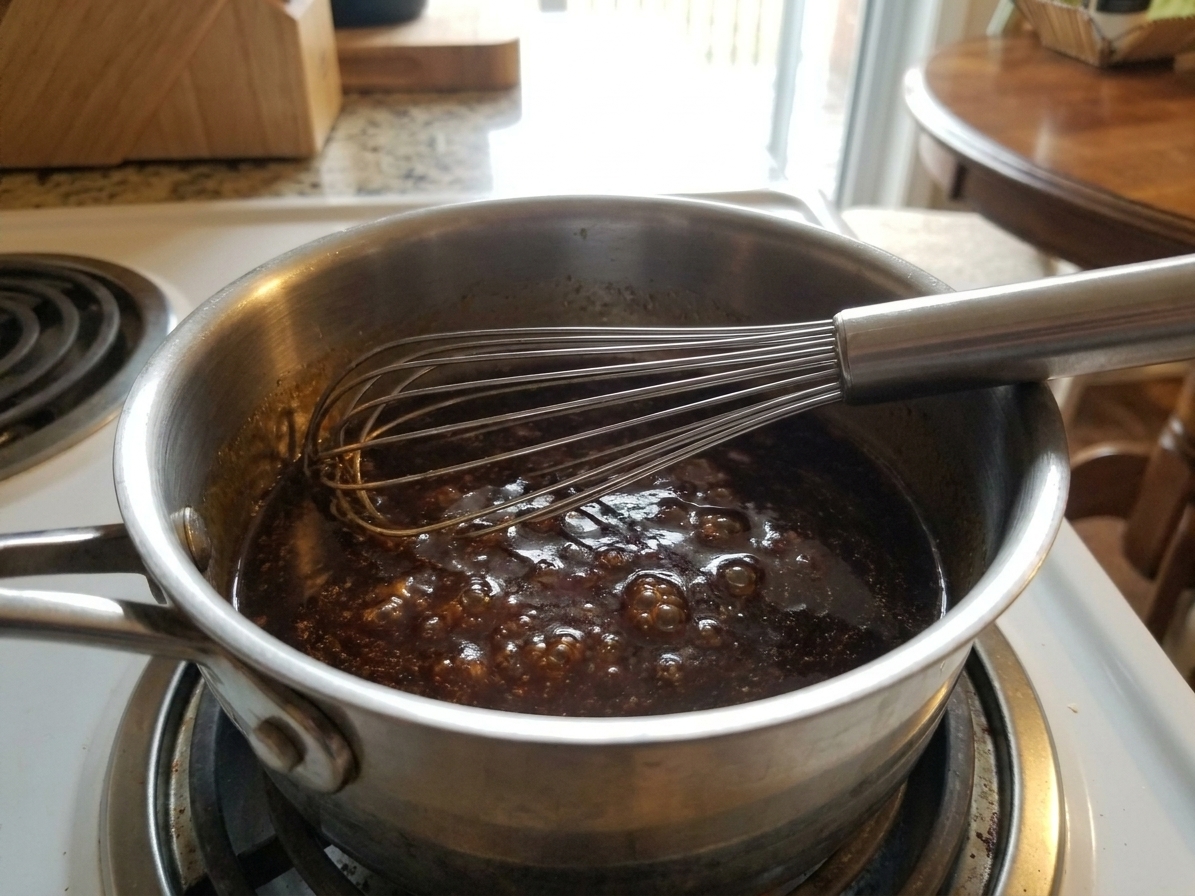 A small saucepan of glossy soy garlic glaze simmering on a stovetop with a whisk resting on the rim