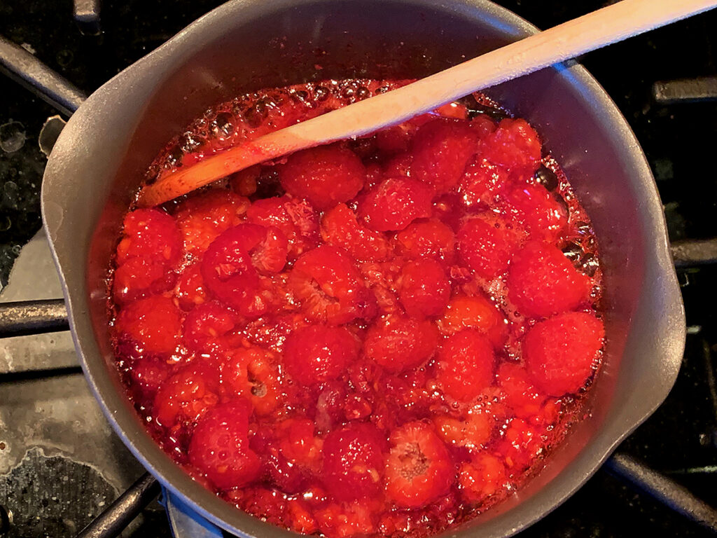 A small saucepan of raspberries simmering into a thick sauce on a stovetop with a wooden spoon resting nearby