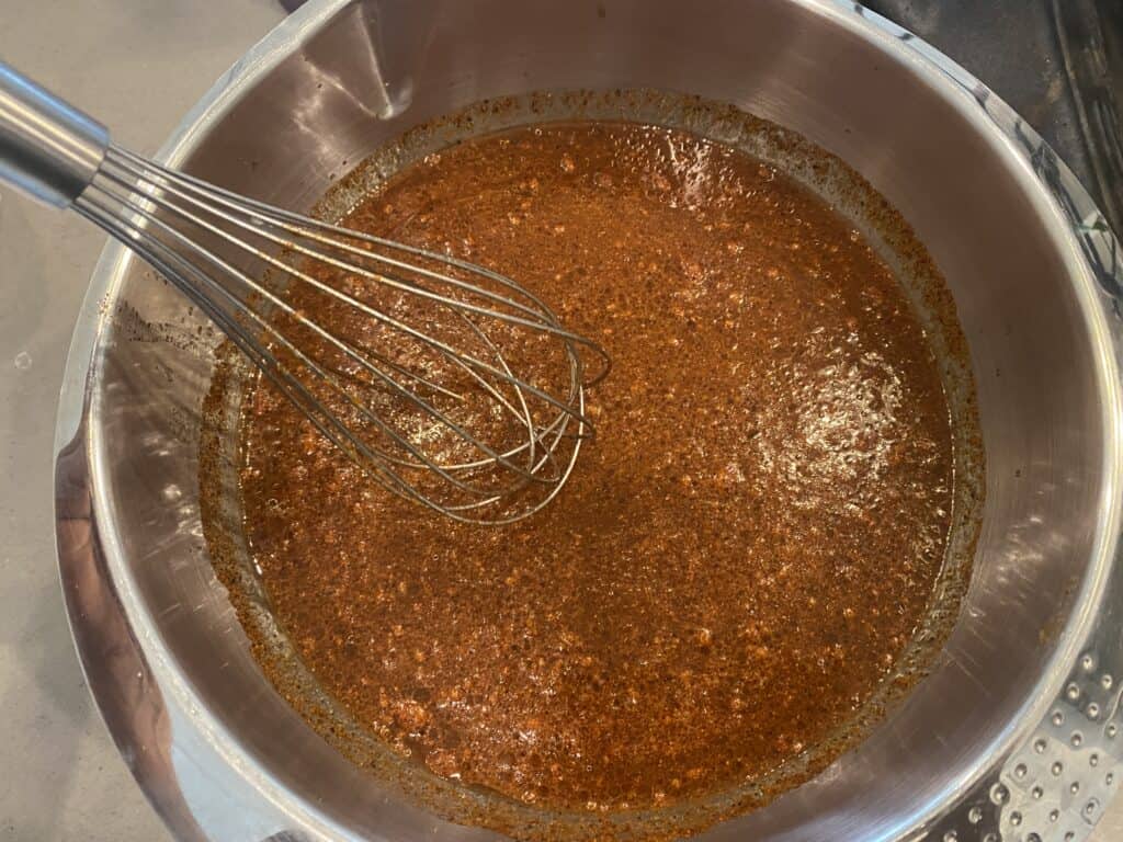 A small saucepan of reddish-brown vinegar mop sauce gently steaming on a stovetop, with a basting brush resting beside it on a light kitchen counter