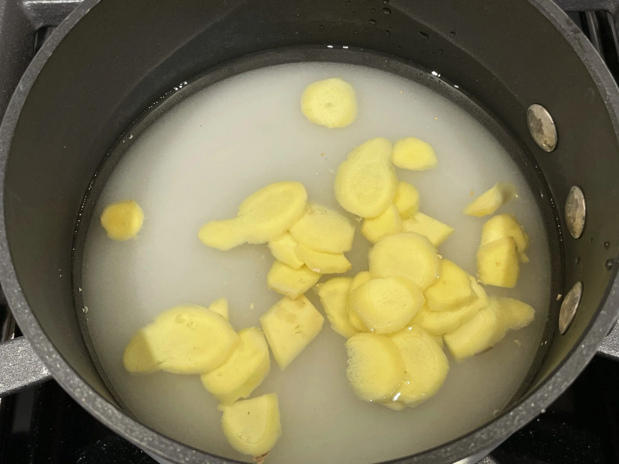 A small saucepan of simmering ginger slices in simple syrup on a home stove with a wooden spoon nearby