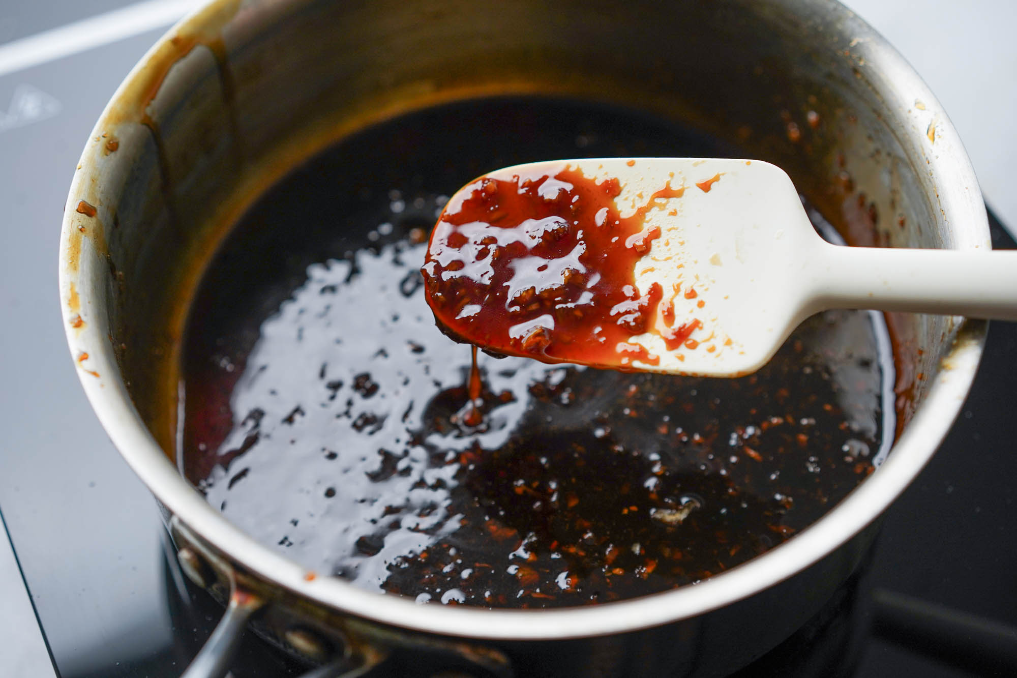 A small saucepan on a stove with a glossy brown sauce simmering, a wooden spoon resting on the rim, warm kitchen lighting