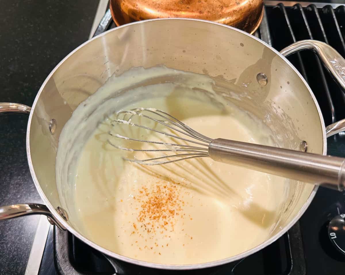 A small saucepan on a stove with creamy Mornay sauce being whisked until smooth, warm kitchen lighting