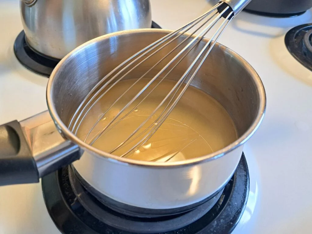 A small saucepan on a stovetop with bubbling honey sugar syrup and a candy thermometer clipped to the side