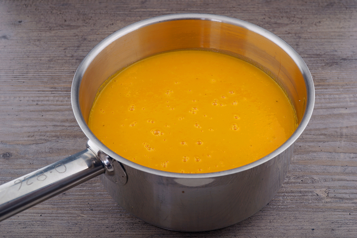 A small saucepan on a stovetop with orange-colored sauce being whisked as it begins to simmer