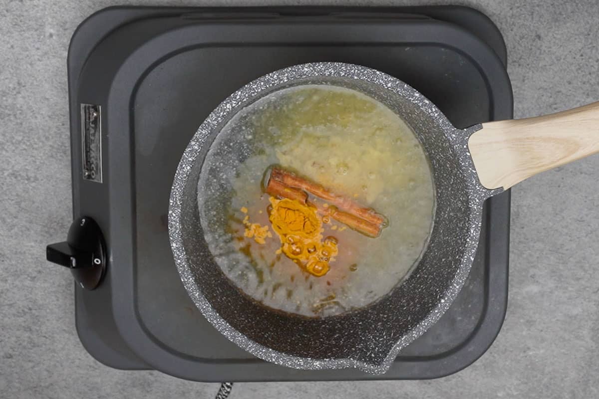 A small saucepan on a stovetop with sliced ginger and a cinnamon stick, with ground turmeric nearby on a spoon