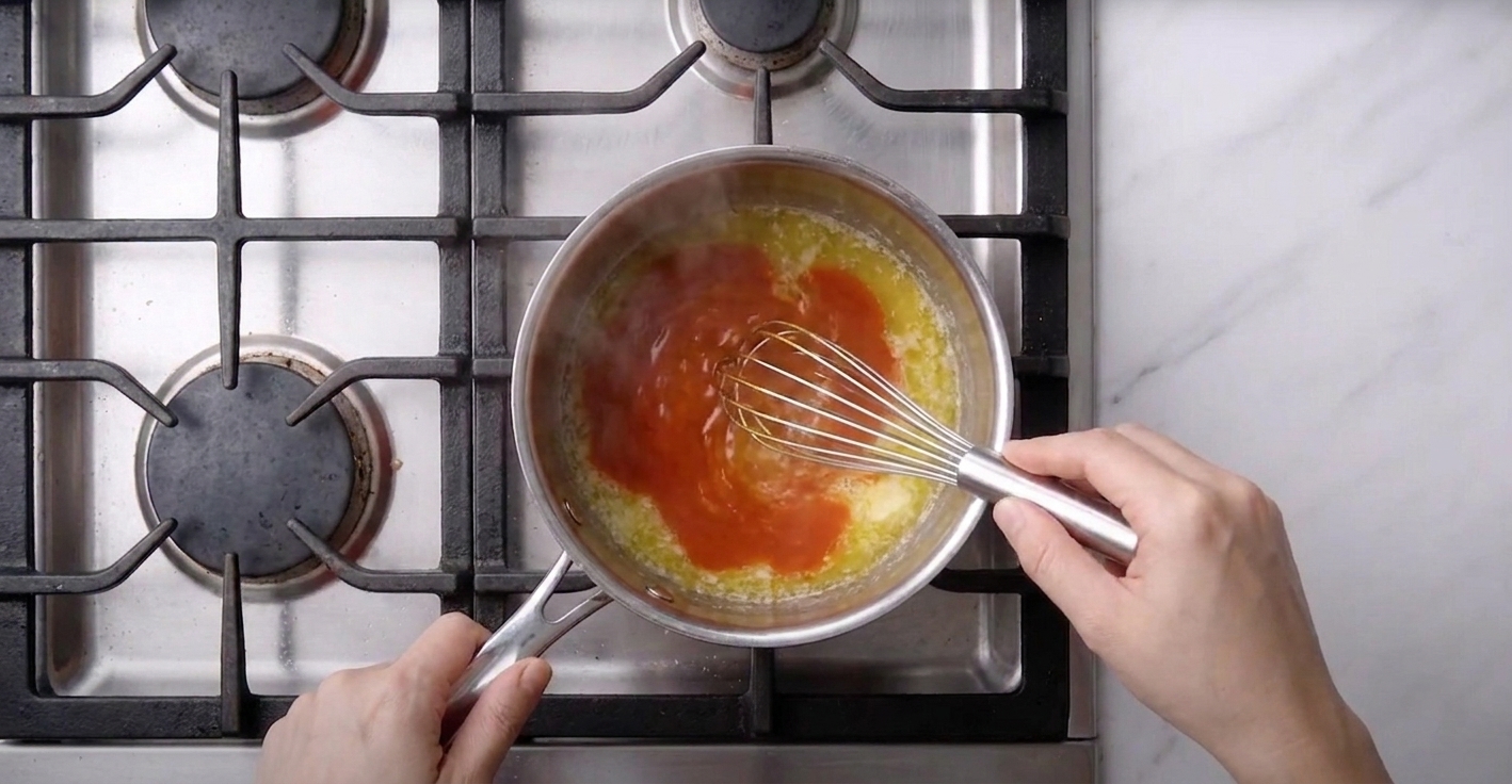 A small saucepan on the stove with melted butter and red hot sauce being whisked together