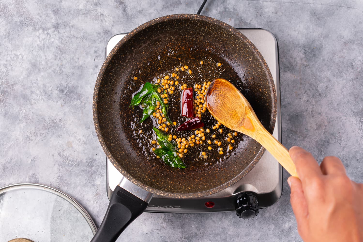 A small skillet on a stovetop with mustard seeds popping in oil alongside curry leaves and dried red chiles