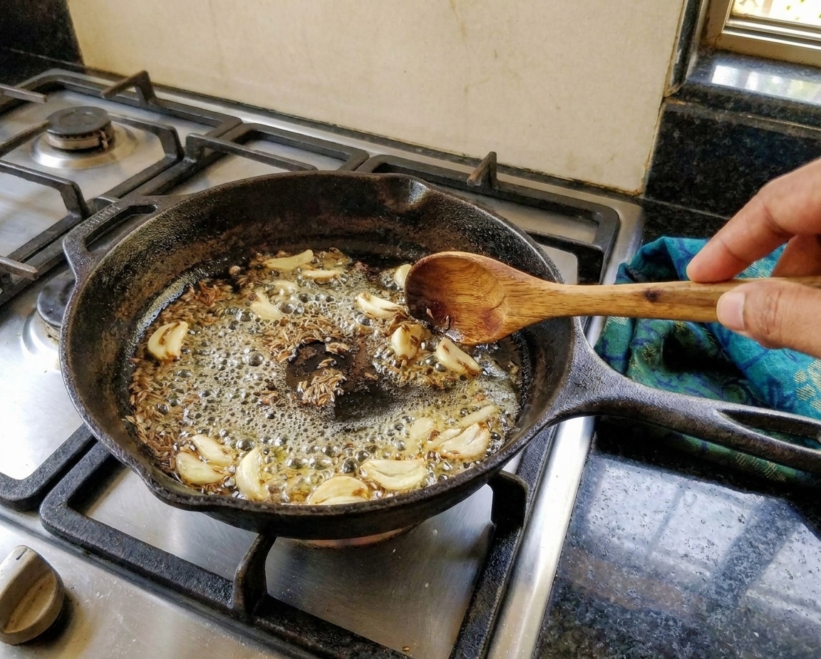 A small skillet with ghee sizzling around cumin seeds and sliced garlic turning lightly golden on a stovetop