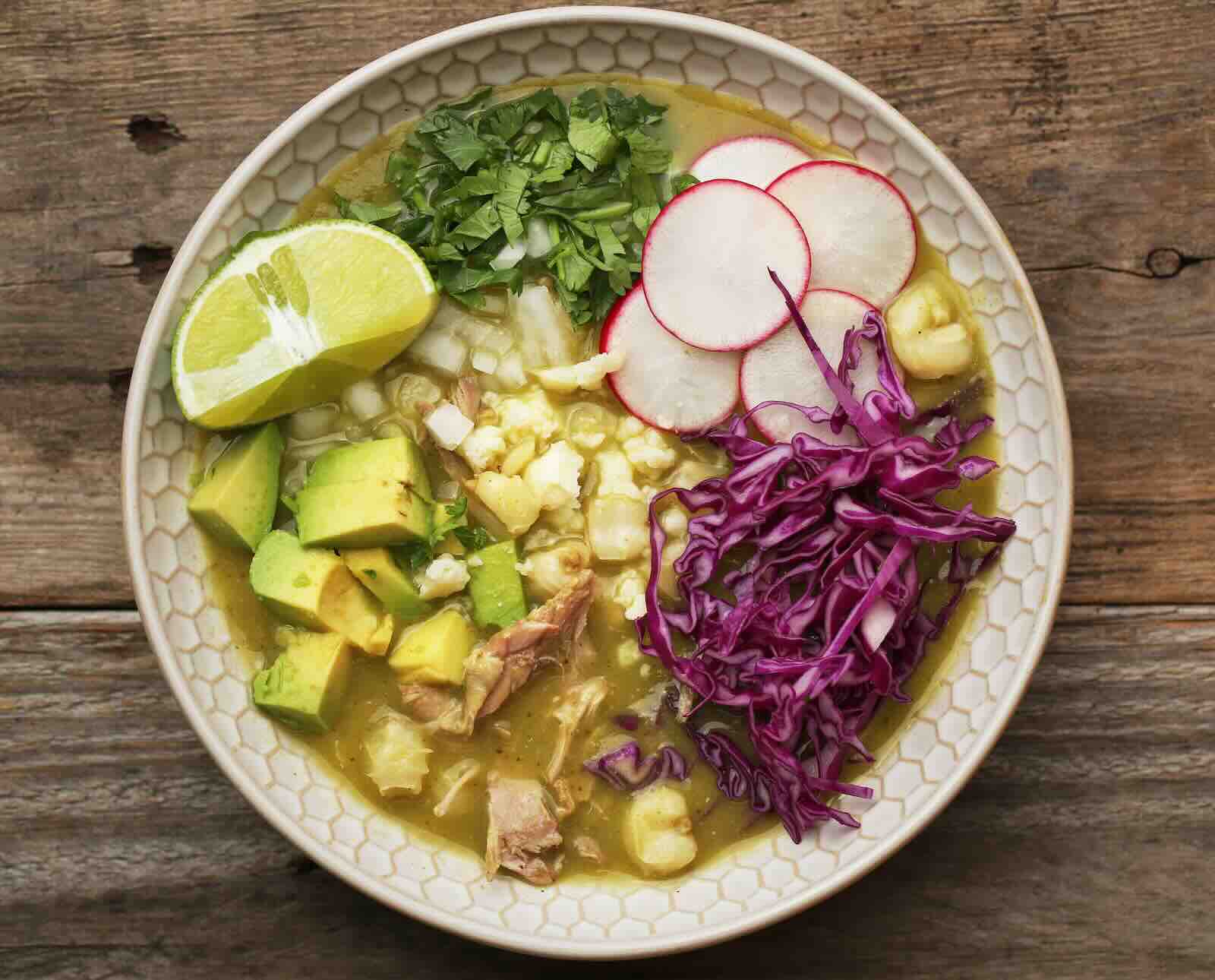 A small spread of pozole toppings including shredded cabbage, sliced radishes, diced onion, chopped cilantro, dried oregano, and lime wedges on a kitchen counter