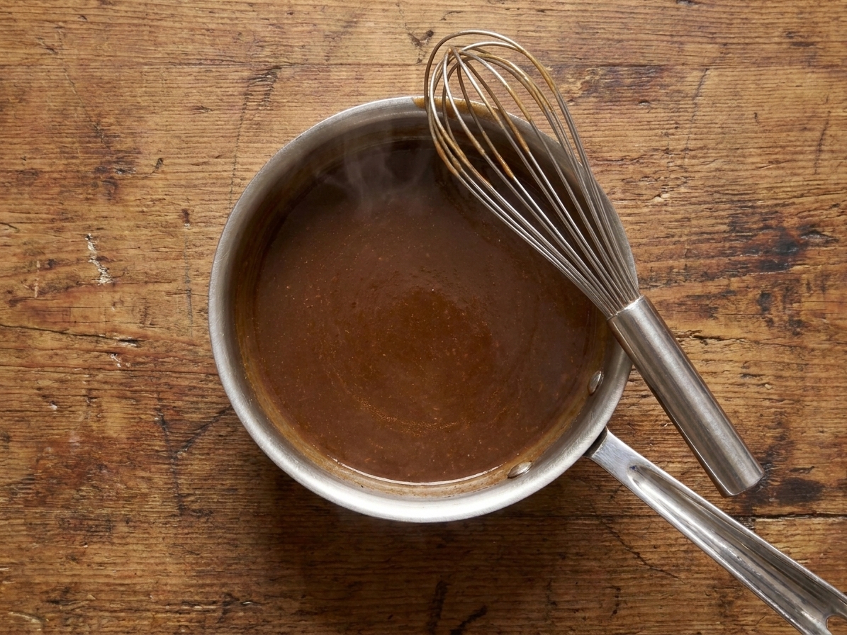 A small stainless saucepan filled with glossy brown au jus gravy, with a whisk resting on the rim on a warm-toned kitchen counter