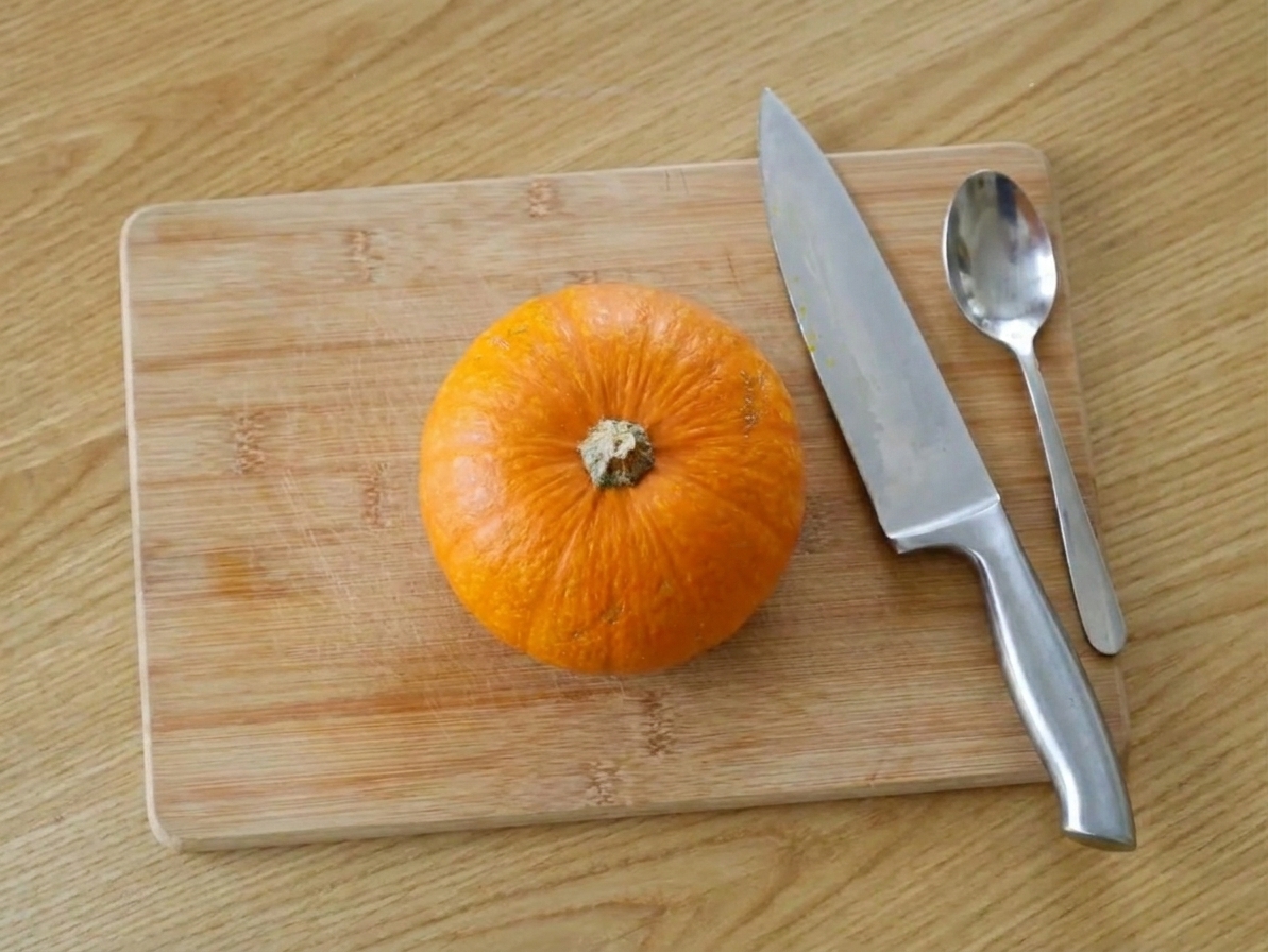 A small sugar pumpkin on a cutting board with a chef's knife and a spoon nearby