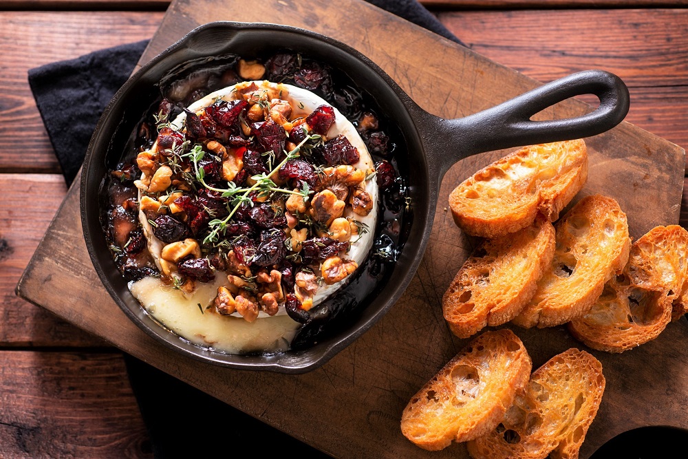 A small wheel of baked Brie bubbling in the center of a cast iron skillet with chopped herbs, sliced garlic, and a drizzle of honey, surrounded by crackers on a warm kitchen counter