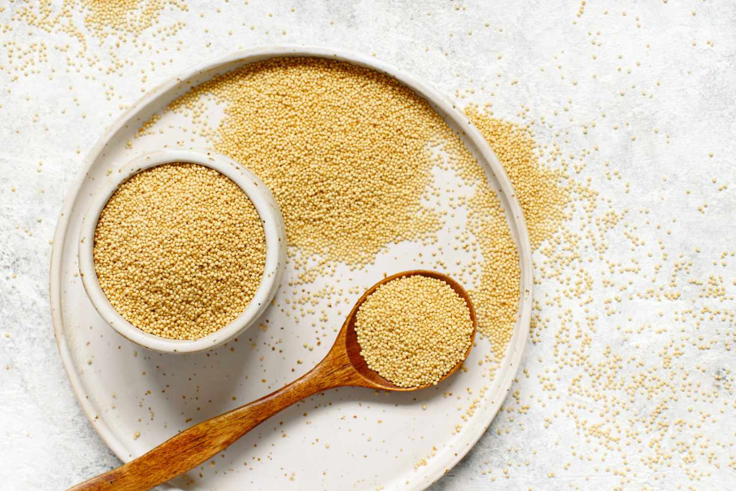 A small white bowl filled with raw amaranth grains on a wooden countertop with a measuring spoon nearby