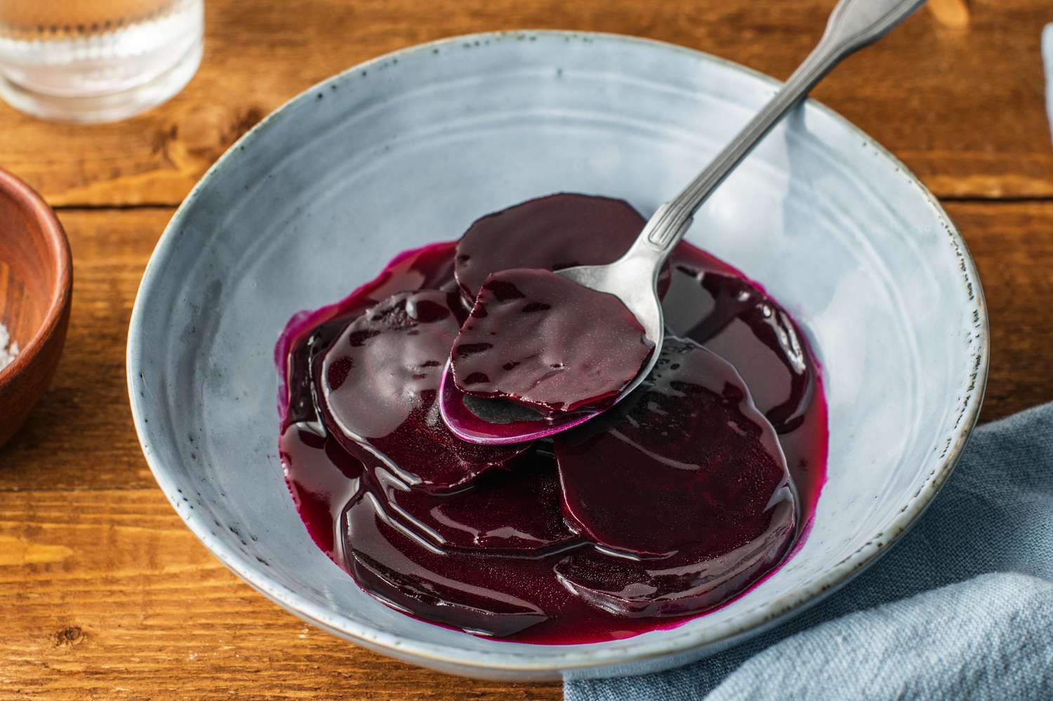 A small white serving bowl filled with Harvard beets in a glossy deep red sweet and tangy sauce, set on a warm kitchen table with a spoon beside it