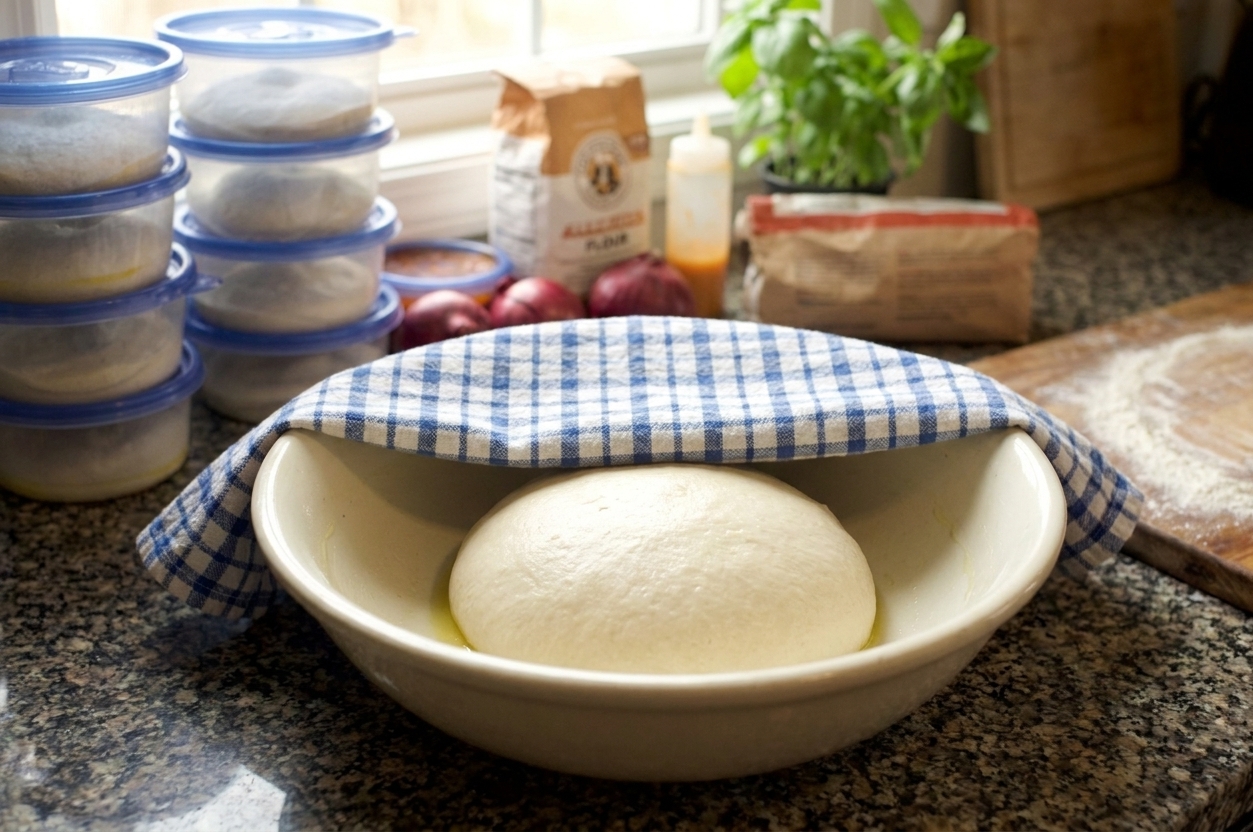A smooth ball of homemade pizza dough resting in a lightly oiled bowl on a warm kitchen counter, covered with a towel