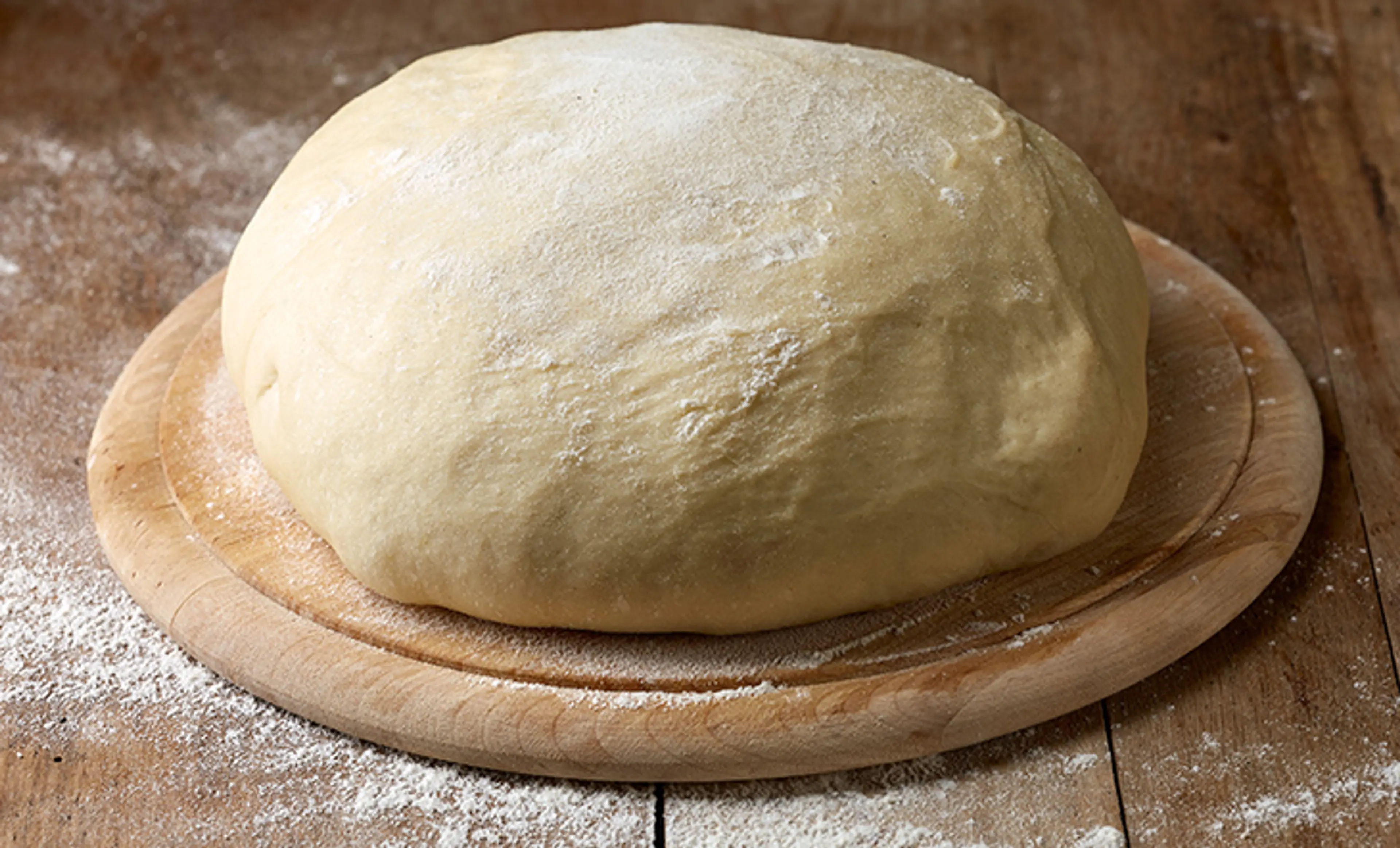A smooth ball of pizza dough resting on a lightly floured kitchen counter with a wooden rolling pin and a small bowl of olive oil nearby