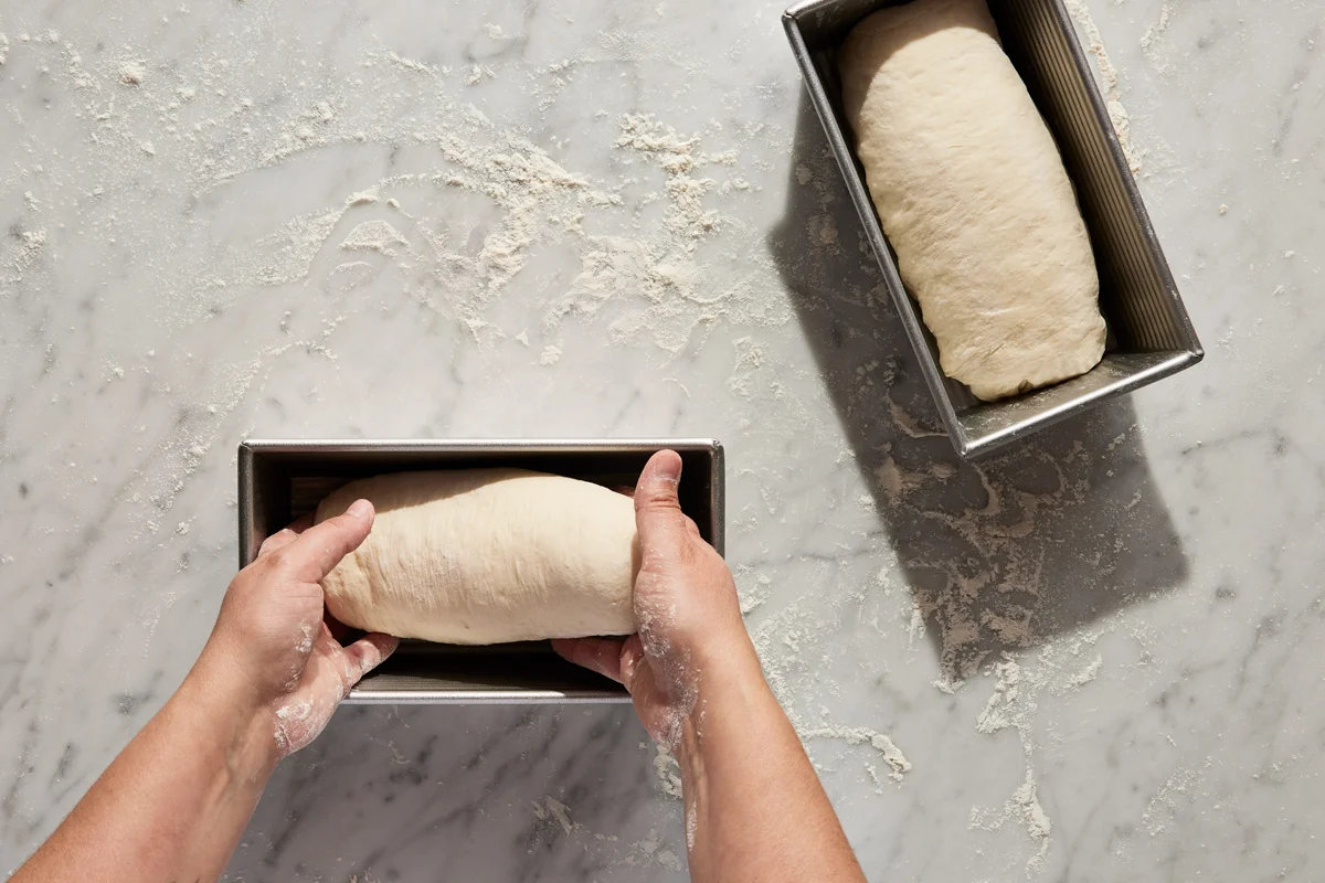 A smooth mound of bread dough domed above the rim of a greased loaf pan, rising on a kitchen counter
