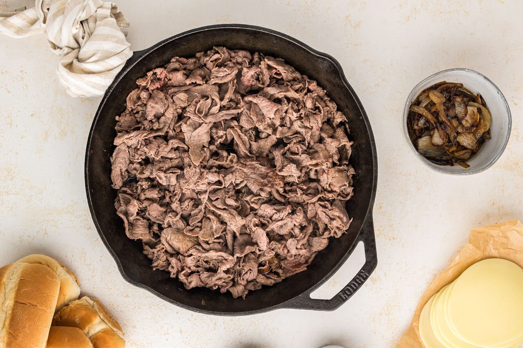 A spatula portioning cheesy shaved steak with onions and peppers into sections in a skillet, ready to load into toasted hoagie rolls