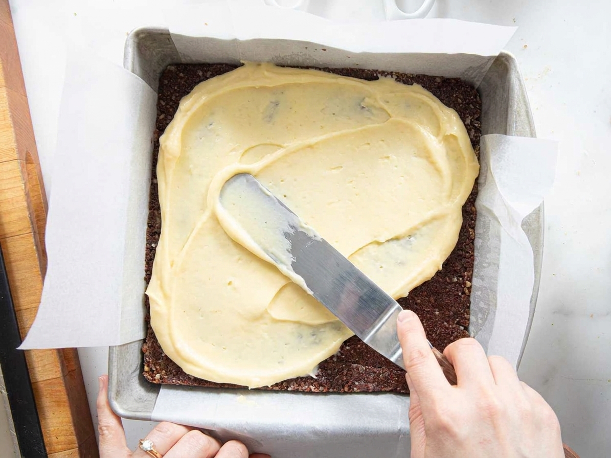 A spatula spreading pale custard buttercream evenly over the chilled chocolate coconut base in a square pan