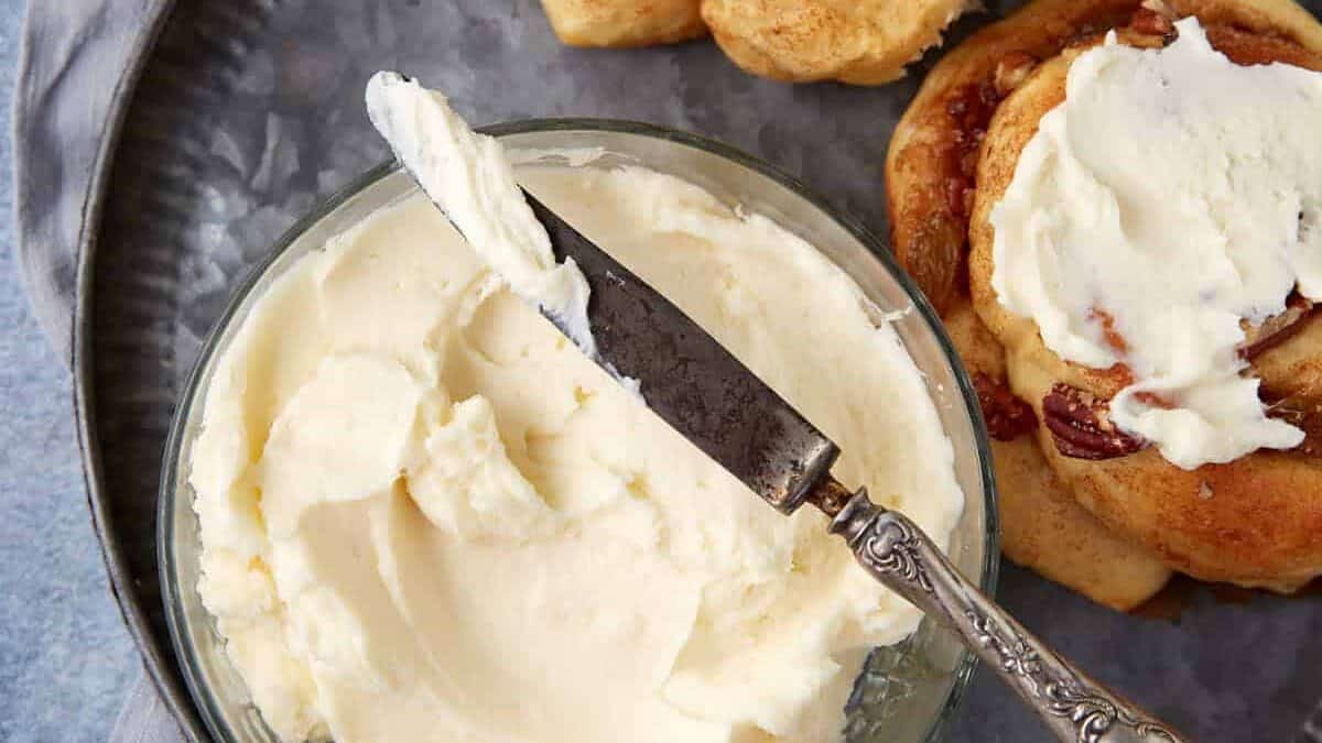A spatula spreading thick cream cheese frosting over warm cinnamon rolls in a baking dish on a bright kitchen counter