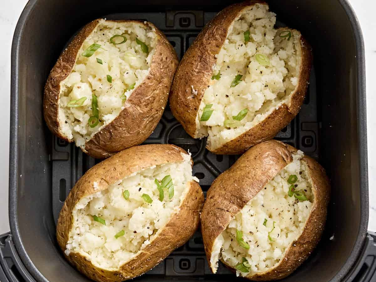 A split air fryer baked potato with crisp, salted skin and fluffy interior, topped with butter and chives on a simple white plate on a warm kitchen table