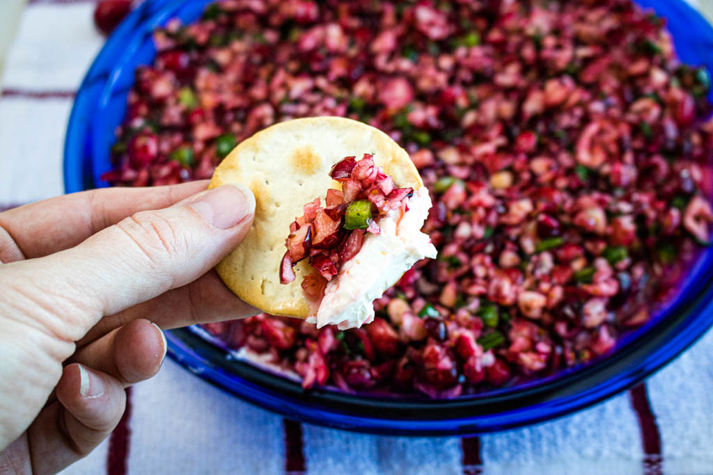 A spoon adding cranberry jalapeño relish on top of a smooth cream cheese base on a serving platter